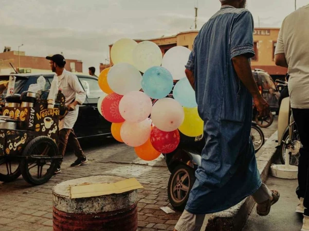 City street scene with an adult male in a blue robe walking past a parked moped laden with colorful balloons, and a coffee vendor.