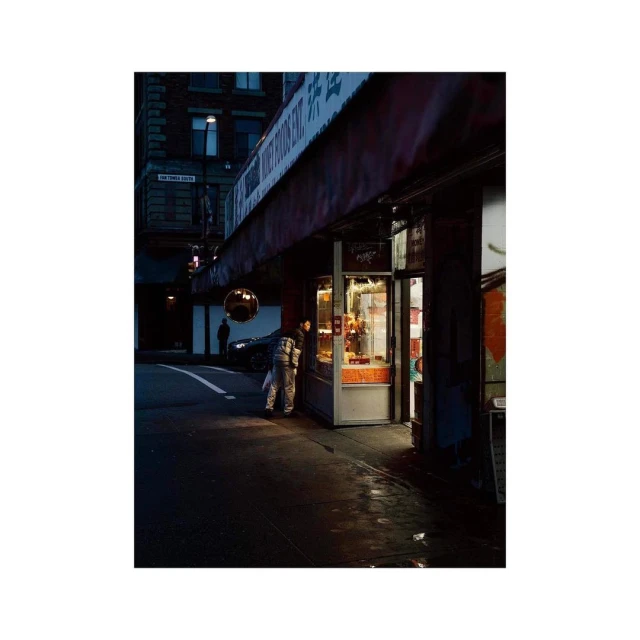 Night scene of two adults looking into a brightly lit food stall in an urban area, street with buildings in the background.