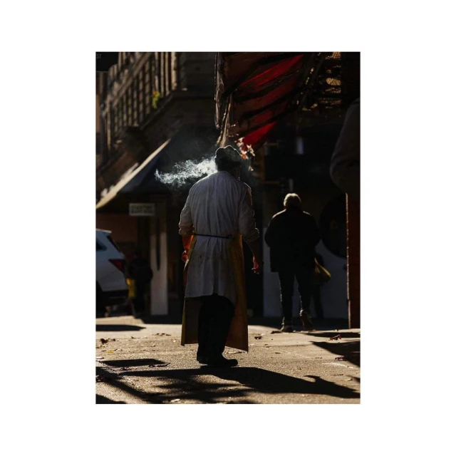 A man exhales cigarette smoke in an urban alleyway, wearing a white lab coat and apron, with pedestrians walking in the background.