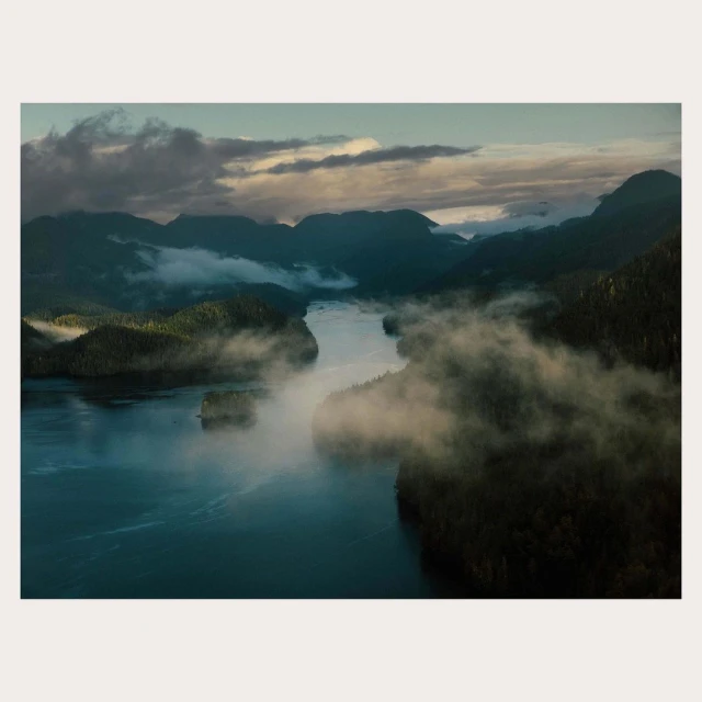 Aerial view of a river cutting through a mountain range shrouded in fog, creating a serene nature scene with lush forests.
