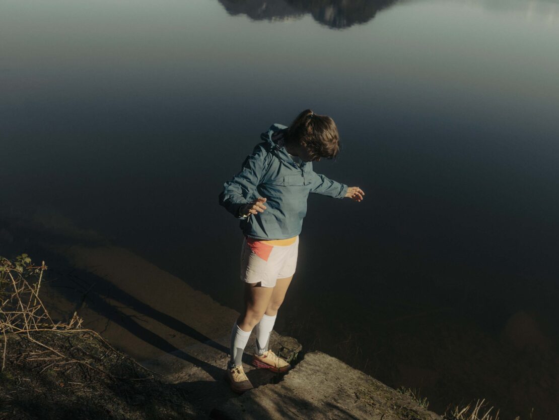 Female figure in jacket and shorts stands on rock near dark lake with distant mountain, bathed in soft, diffused sunlight.