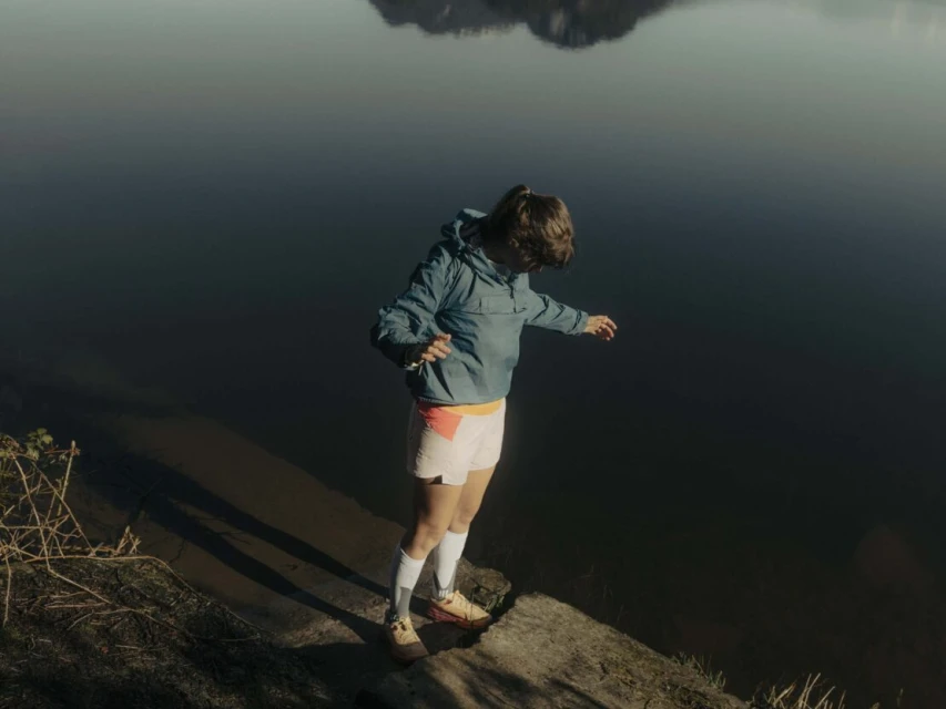 Female figure in jacket and shorts stands on rock near dark lake with distant mountain, bathed in soft, diffused sunlight.