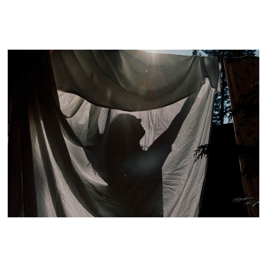 Silhouette of a person holding up a translucent white cloth outdoors against a backdrop of sunlit trees, creating an ethereal scene.