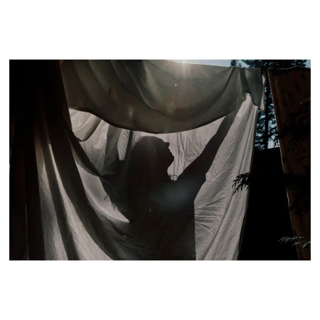 Silhouette of a person holding up a translucent white cloth outdoors against a backdrop of sunlit trees, creating an ethereal scene.