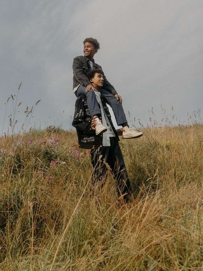 Two young men in a grassy field, one carrying the other on his shoulders under a cloudy sky, enjoying an outdoor activity.