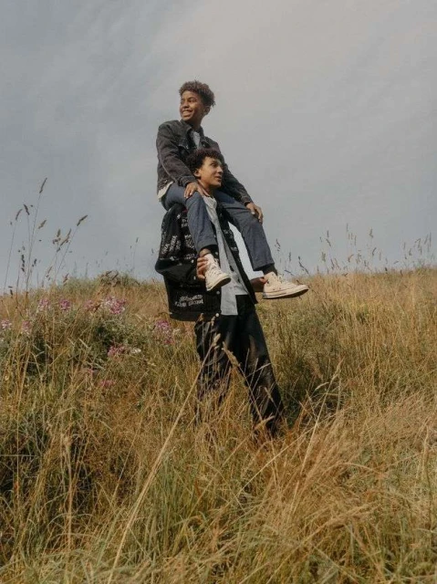 Two young men in a grassy field, one carrying the other on his shoulders under a cloudy sky, enjoying an outdoor activity.