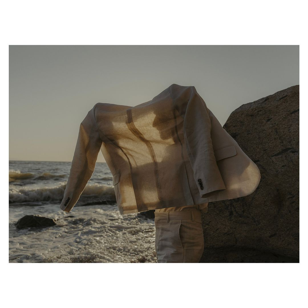 Beige linen suit jacket and pants arranged on a rock at the seashore with the ocean and sky visible in the background.