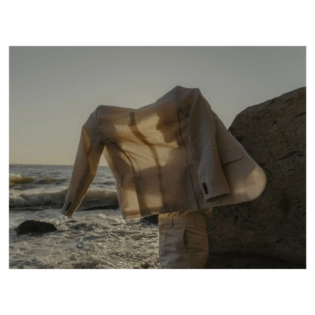 Beige linen suit jacket and pants arranged on a rock at the seashore with the ocean and sky visible in the background.