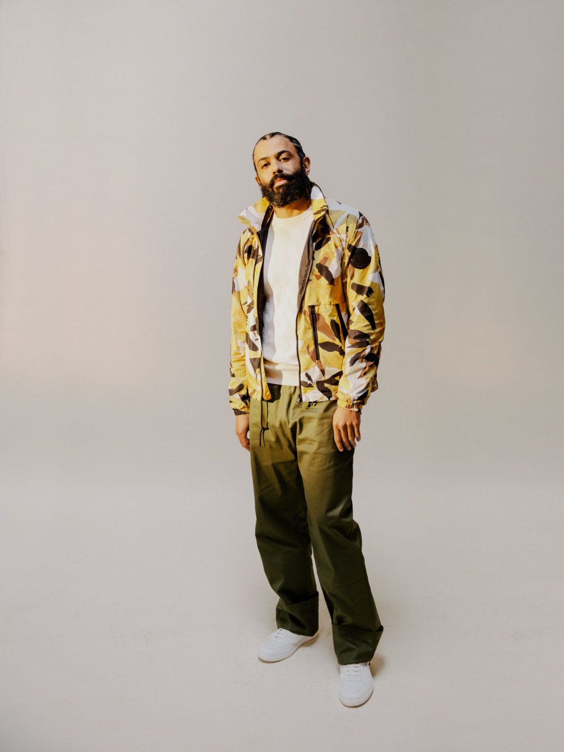 An adult male model poses indoors wearing a green coat, white shirt, and brown pants, with hair slicked back, in a portrait shot.