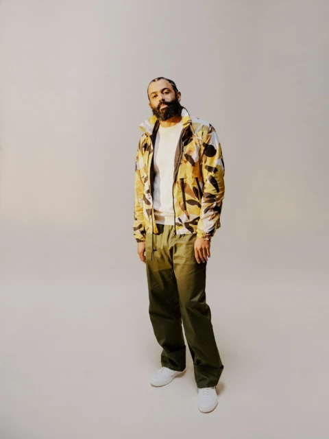 An adult male model poses indoors wearing a green coat, white shirt, and brown pants, with hair slicked back, in a portrait shot.