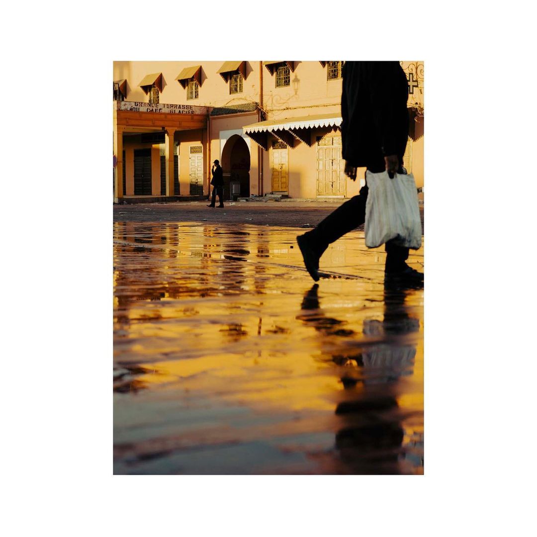 Person walking through wet, sunlit Moroccan street, casting reflection on the ground; light bounces off buildings.
