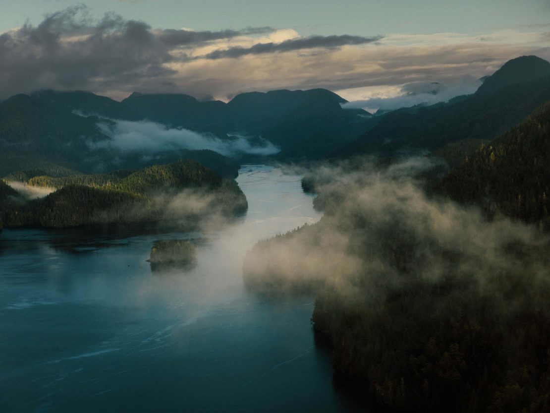 Aerial view of a body of water surrounded by forest-covered mountains, shrouded in mist and illuminated by soft, natural lighting.
