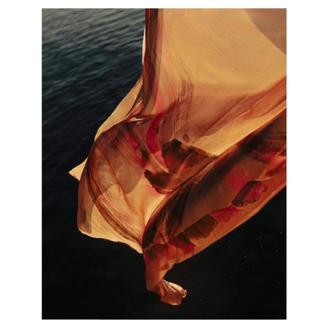 Abstract studio photograph featuring a flowing beige textile with blurred red and brown patterns against a dark blue water backdrop.