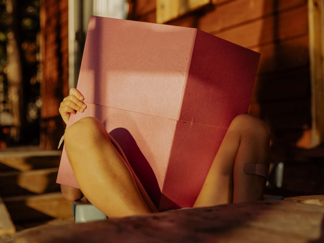 Child sits with large pink cube covering face, outdoors with wooden background. Bandage on leg, golden hour lighting.