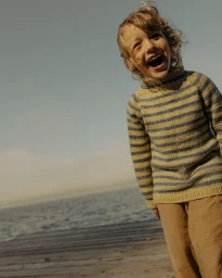 A laughing child with curly hair wearing a striped turtleneck sweater on a wooden dock overlooking water.