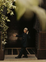 Elderly man in a dark suit gesturing gracefully inside a vaulted doorway with ornate tile patterns