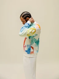 Man with braided hair wearing a white watercolor-printed sweater and white trousers against a neutral backdrop