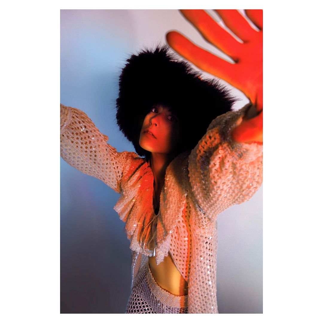 Woman in a sequin dress with a black furry hat extends her hand towards the camera in a studio setting with colorful lighting.