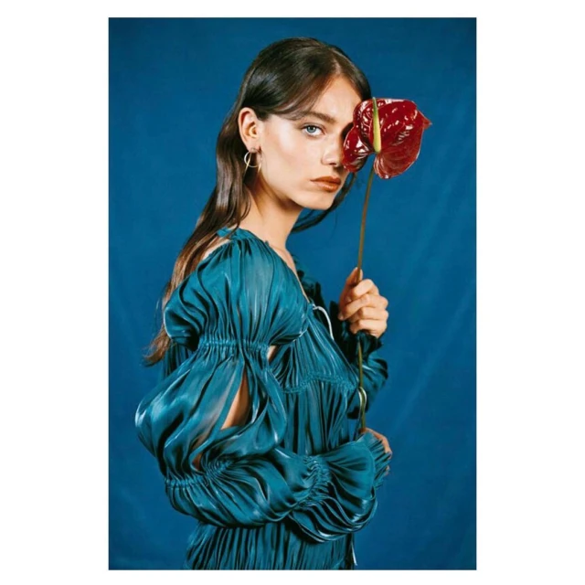 A brunette woman in a teal dress poses against a blue background, holding a red anthurium flower covering part of her face.