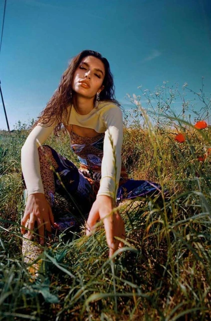 Woman with long dark hair sits in a field with wildflowers under a bright blue sky, wearing a patterned dress and light sweater.