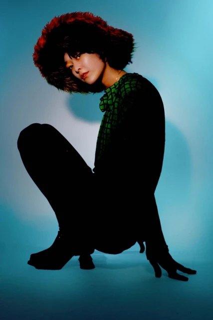 Portrait of an Asian woman with dark hair and a fur hat, squatting in a studio setting with a light blue background and soft lighting.