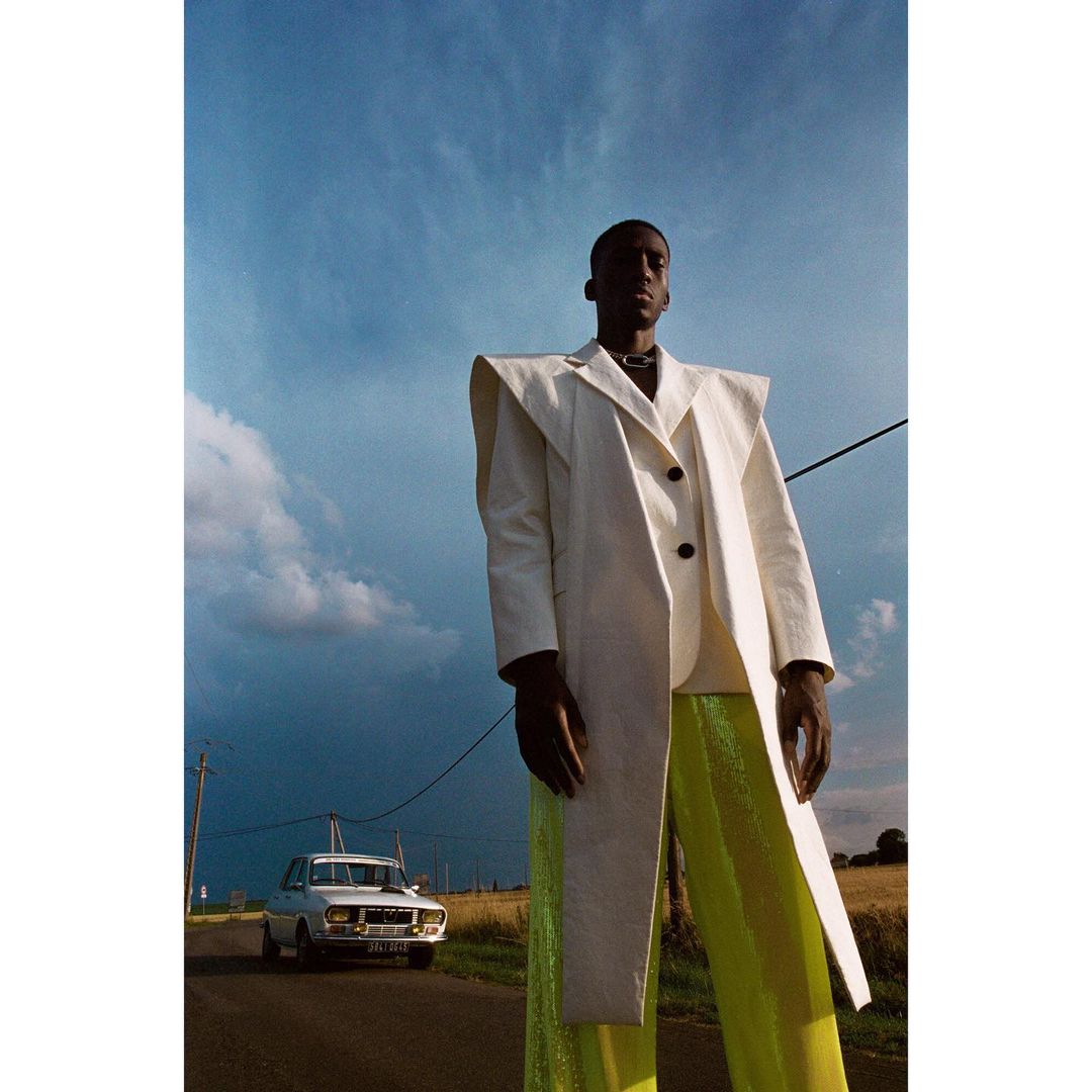 Fashion model in a white coat and shimmering green pants poses on a rural road against a blue sky with a vintage car in the background.