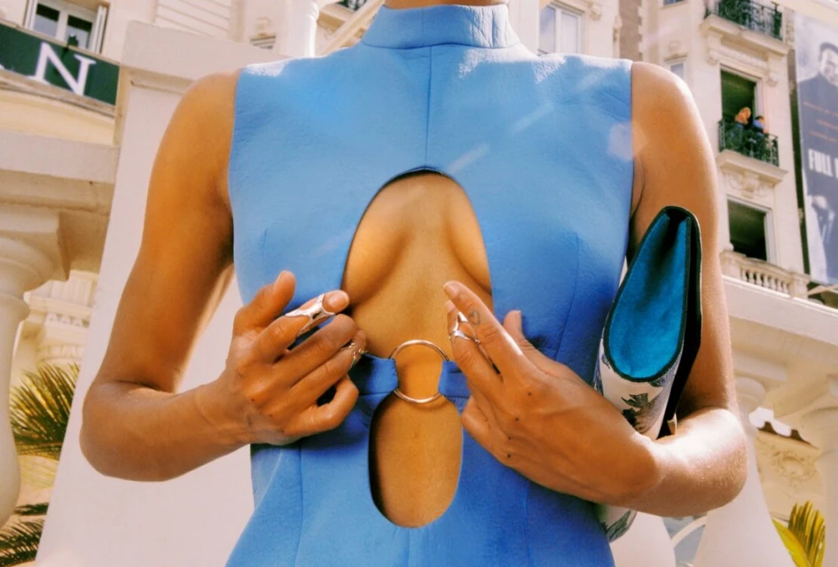 Woman in a blue dress poses outdoors holding a patterned clutch. Close up shot with a building and people in the background.