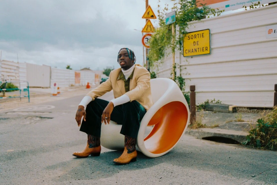 Stylish man sits on modern egg chair on city street, wearing beige jacket, green pants, cowboy boots, under cloudy sky. 