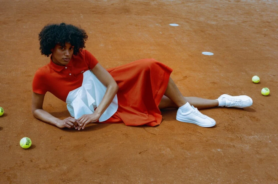 Woman with afro lies on tennis court in orange dress and white sneakers surrounded by tennis balls in medium, outdoor shot.