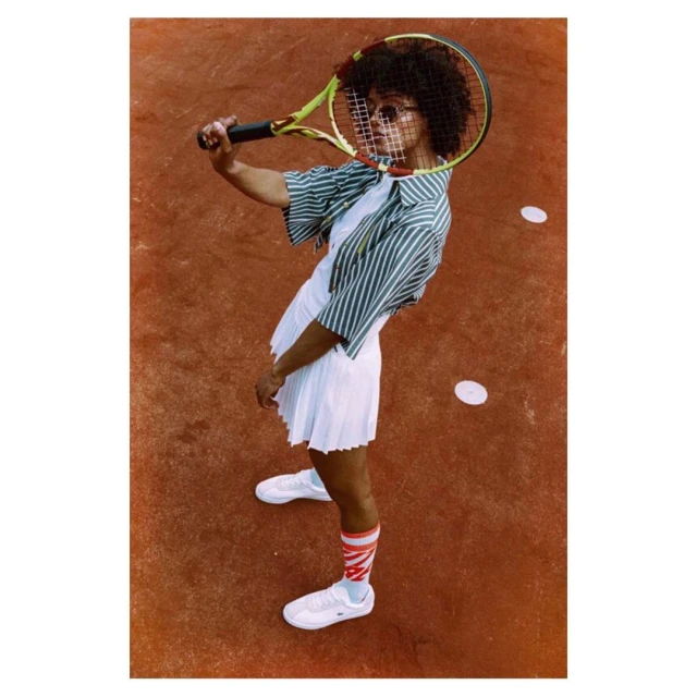 A tennis player with an afro and sunglasses poses on a clay court, holding a racket over her head, wearing a striped shirt and white tennis outfit.