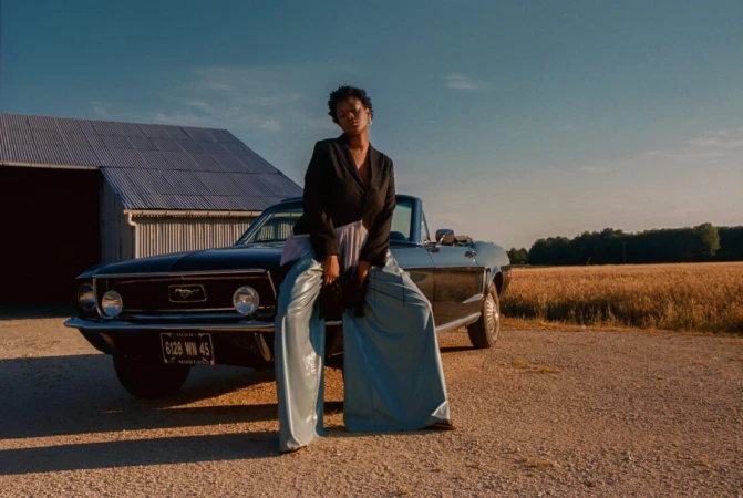 A woman in a black jacket and wide blue pants poses on a blue convertible car in a rural setting with a barn and wheat field.