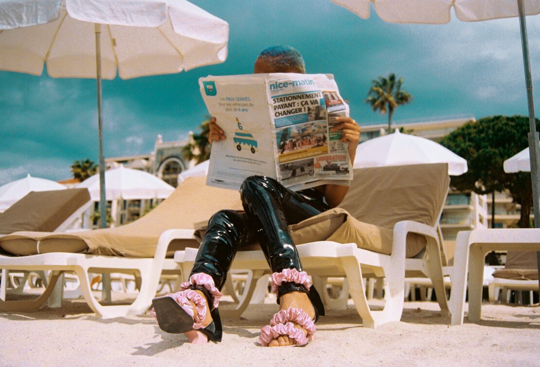 Person reading a newspaper on a beach lounger wearing shiny black trousers and ruffled pink sandals.