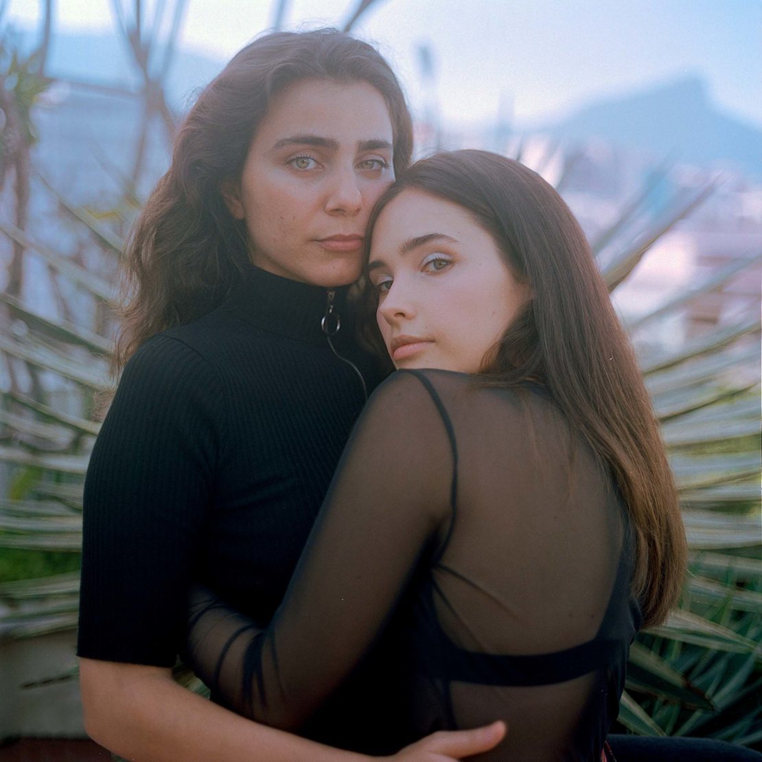 Two women embrace outdoors with soft lighting and a blurry landscape background, showcasing a close, tender moment in a natural setting.