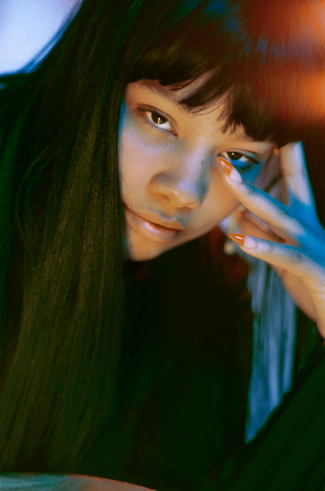 Close-up portrait of a young woman with straight black hair, light skin, and painted nails, set against a dark background with colored lighting.