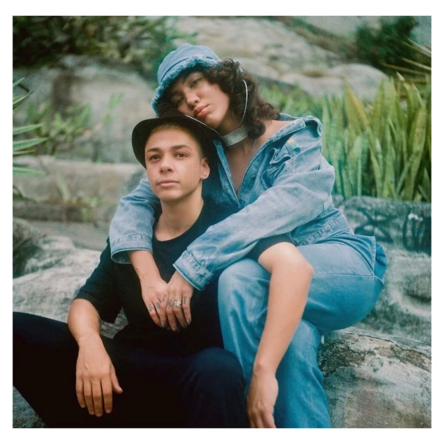 Two people, one sitting and one leaning behind, pose on a rock formation with greenery in the background, both in denim and black attire.