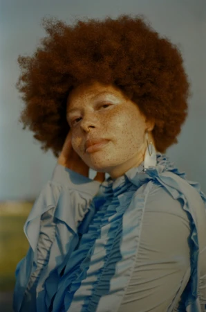 Portrait of a black woman with a large afro hairstyle and freckles wearing a light blue ruffled blouse by Kamila K Stanley.