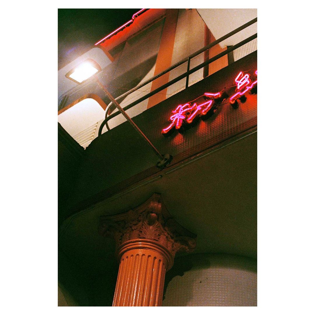 Low-angle shot showcases a column supporting an overhang, bathed in the warm glow of neon signage and artificial lighting at night.