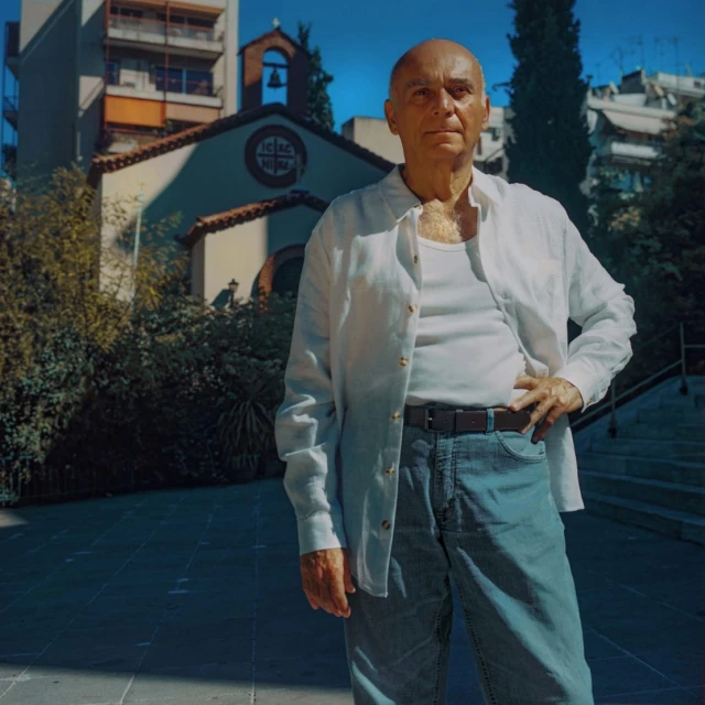 Elderly man stands in an urban setting near a small chapel, wearing a white shirt, tank top, and jeans under a bright blue sky.
