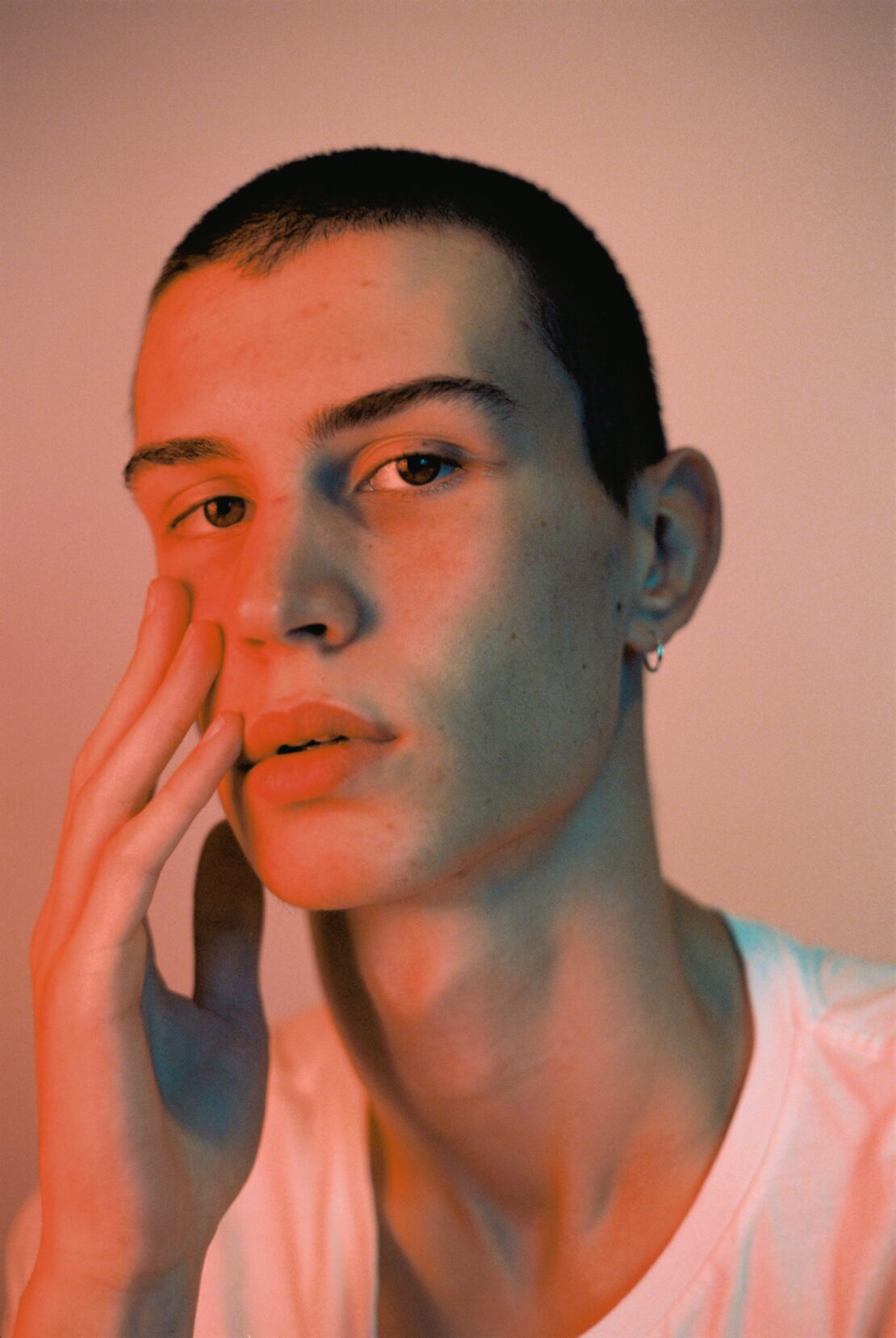 Close-up portrait of a young adult with a buzzed haircut in a white t-shirt, holding their hand to their face in a softly lit studio.