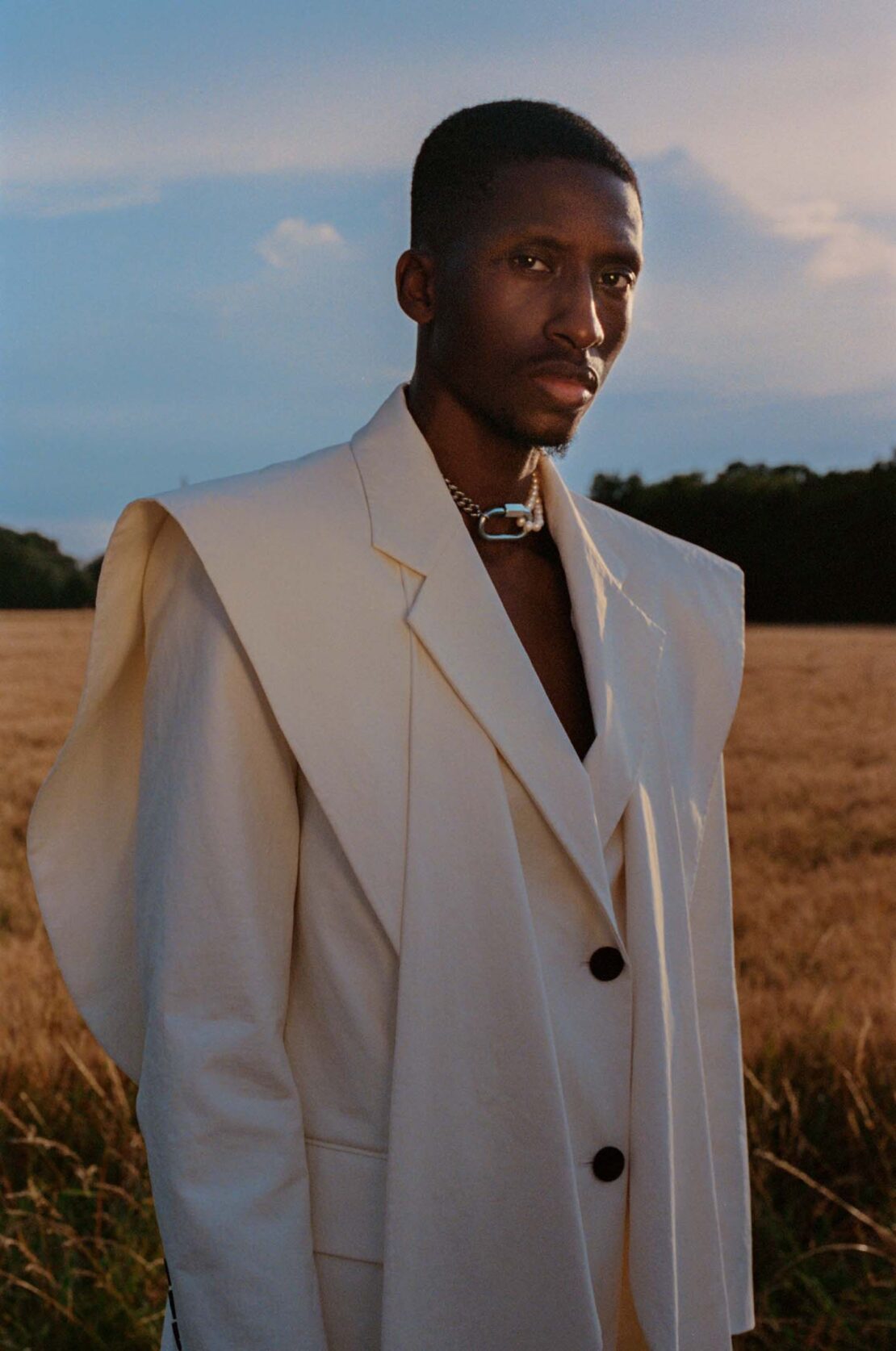 Portrait of an adult Black male model outdoors in wheat field, wearing an oversized cream-colored blazer and chain necklace.