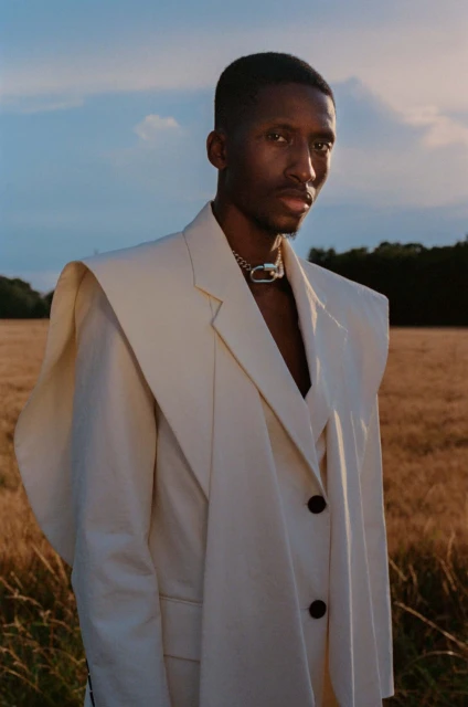 Portrait of an adult Black male model outdoors in wheat field, wearing an oversized cream-colored blazer and chain necklace.