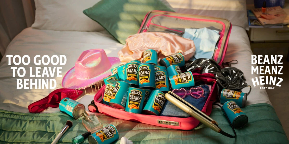 Promotional image of a pink suitcase filled with Heinz Beanz cans, surrounded by travel items on a bed with the tagline 'Too Good to Leave Behind'.