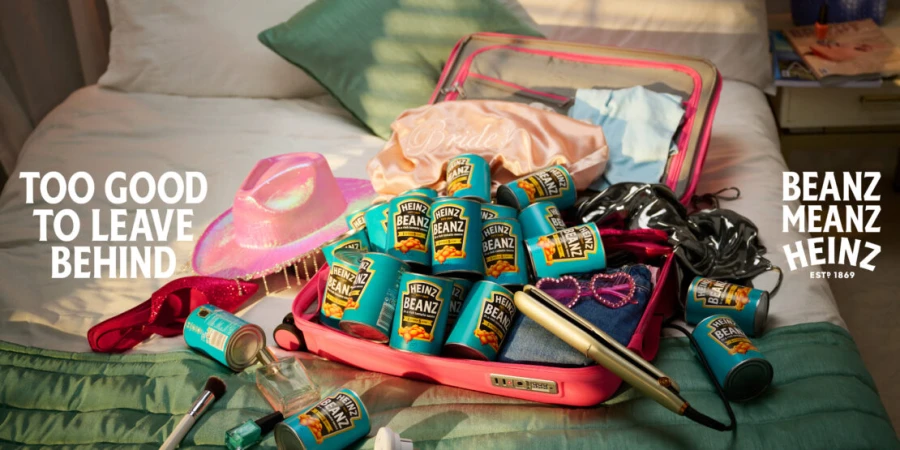 Promotional image of a pink suitcase filled with Heinz Beanz cans, surrounded by travel items on a bed with the tagline 'Too Good to Leave Behind'.