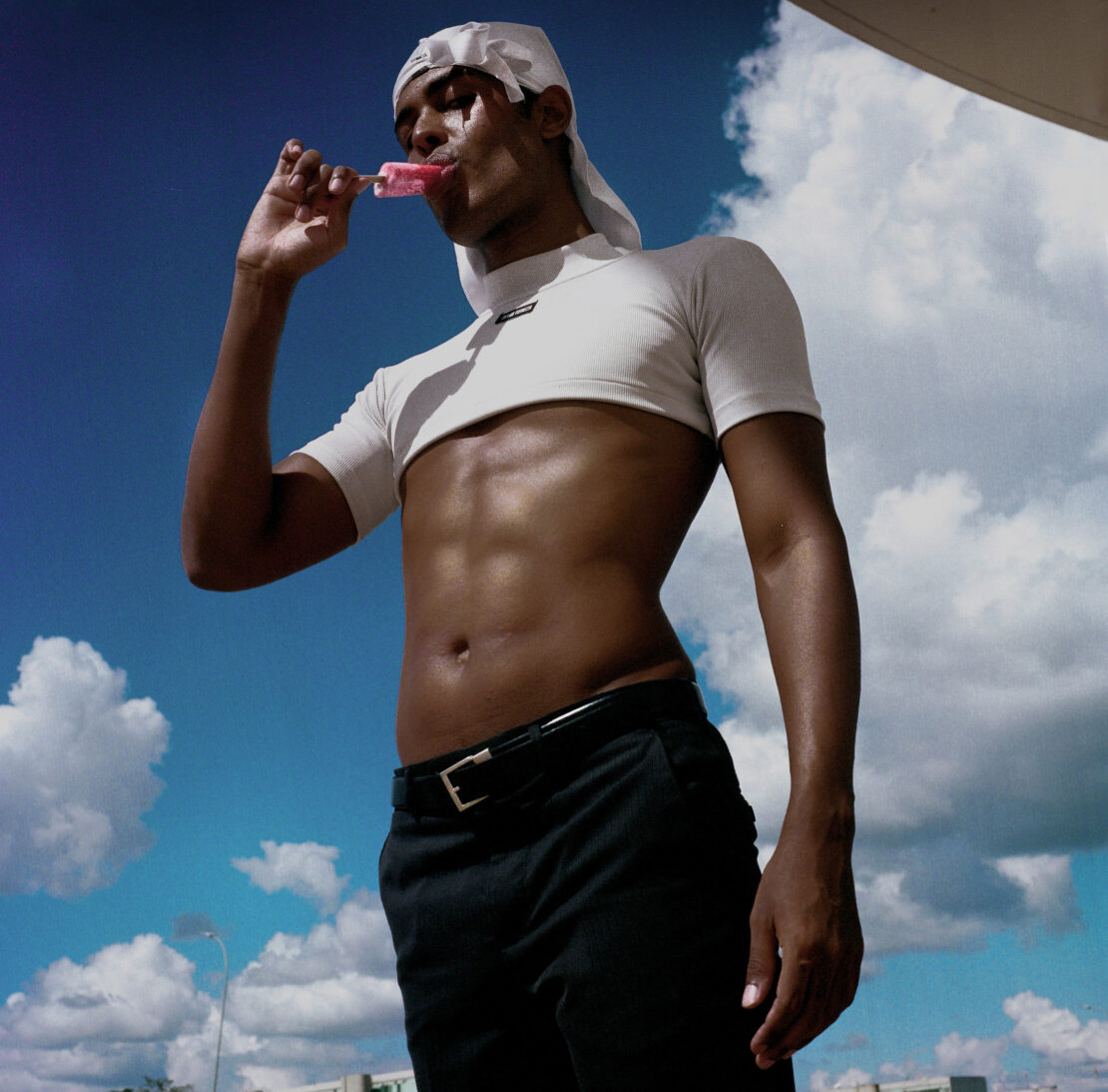 Young adult male with a bare torso, wearing a cropped top and a white headscarf, enjoying a popsicle with a cloudy blue sky background.