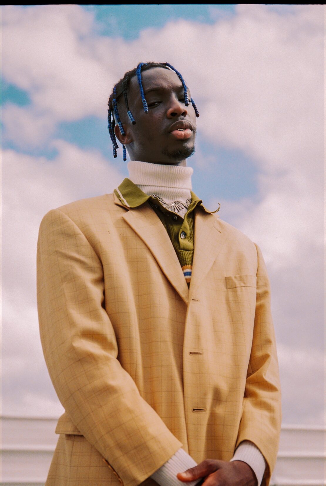 Portrait of a young Black man wearing a tan suit and blue braids, posing against a background of soft blue sky and clouds.