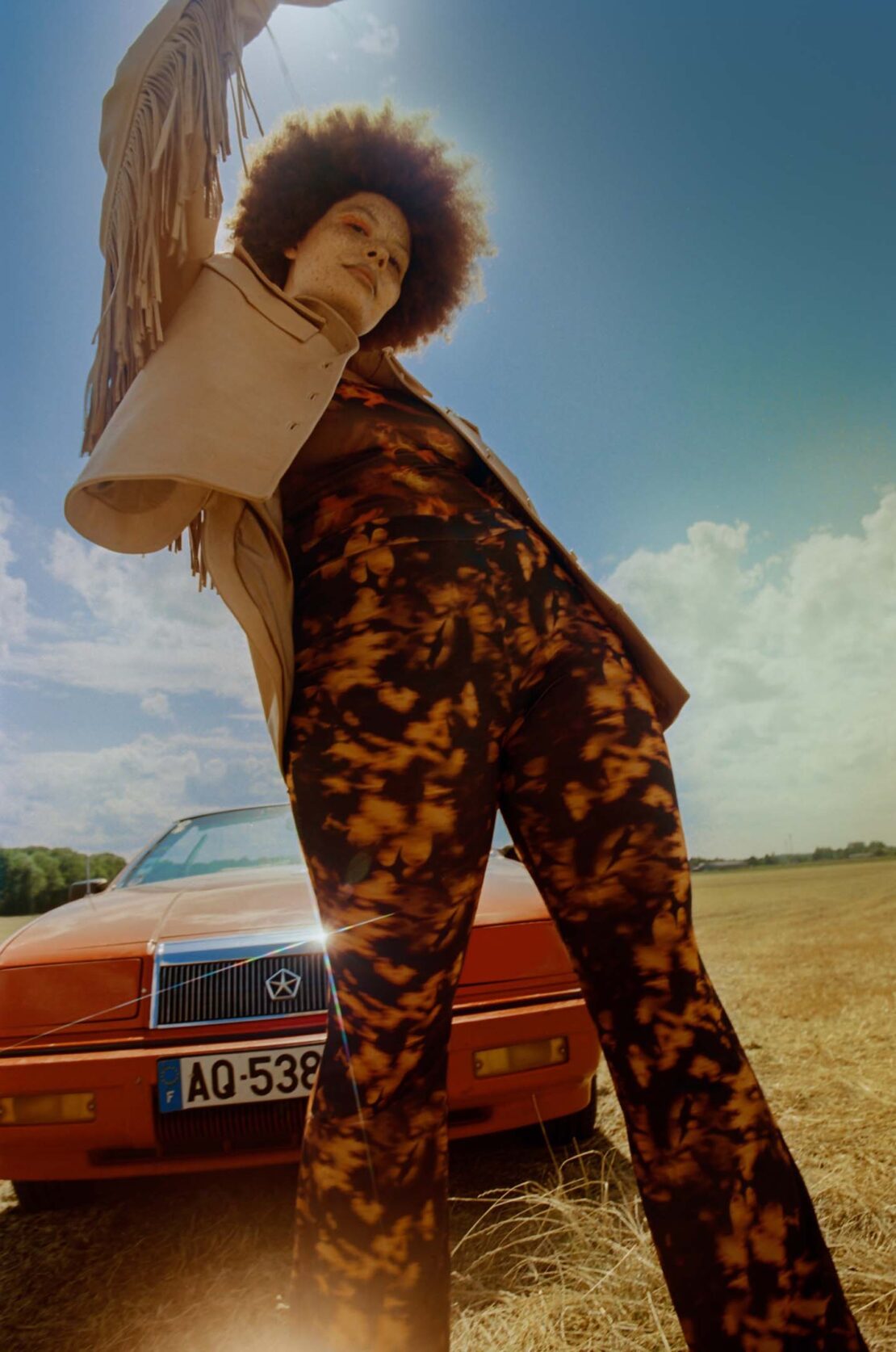 Low-angle portrait of a woman with afro hair wearing a fringe jacket and patterned trousers standing before a red car