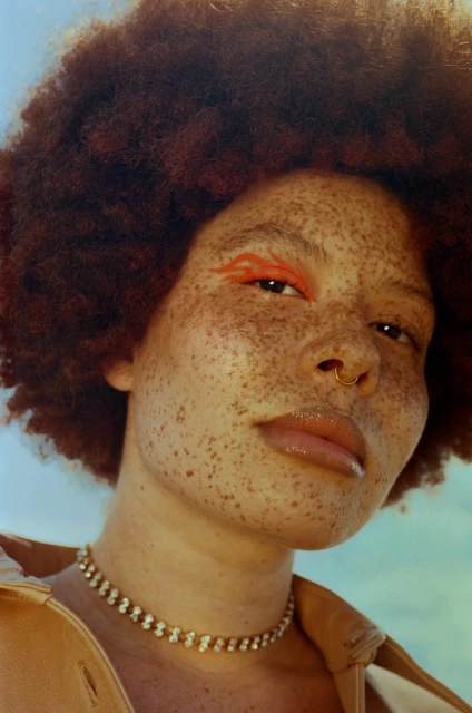 Close-up portrait of a freckled adult female with an afro hairstyle, wearing a gold choker and a tan jacket against a blue background.