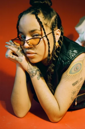 Portrait of a woman wearing cat-eye sunglasses and braids, posing against a solid red backdrop by photographer Kamila K Stanley.
