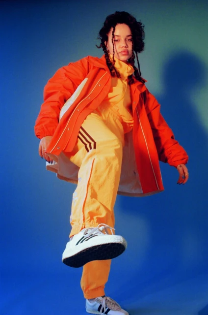 A woman poses in a photo studio with hands on her head wearing a black top and pants, with pink hair and smokey eye makeup.