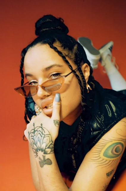Portrait of a woman with sunglasses and braided hair lying down on a red background, showcasing tattoos on her arm and hand.
